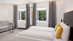 A modern hotel room with a large bed, a sofa, and two windows with curtains. - Stadthotel Oberndorf - 24h Self Check-In Terminal  (Oberndorf bei Salzburg)