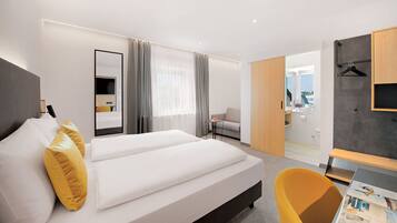 A modern hotel room with a large bed, a small table, a chair, a wardrobe, and a bathroom visible through an open door.