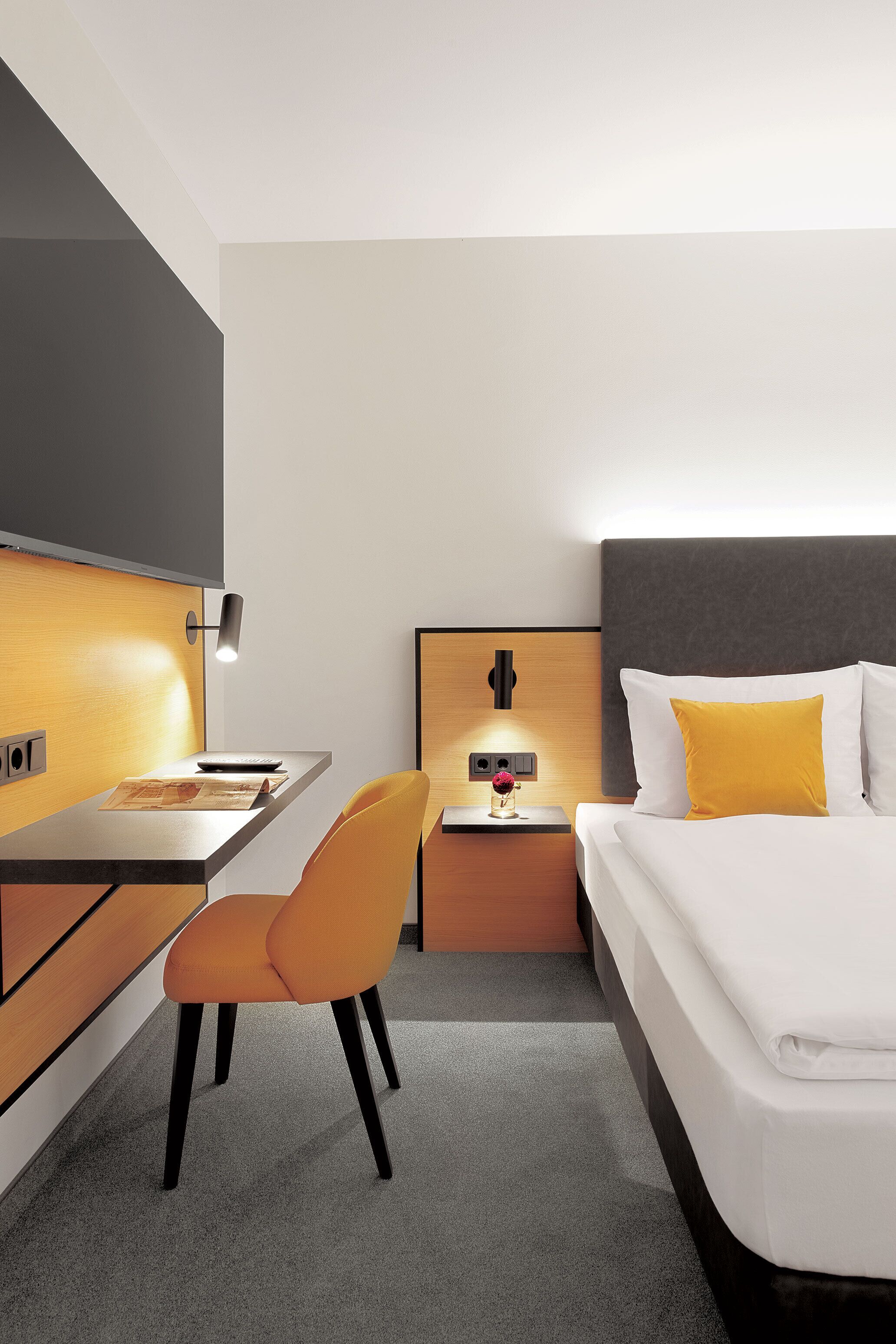 A hotel room with a bed, a desk, a chair, and a nightstand with a lamp.