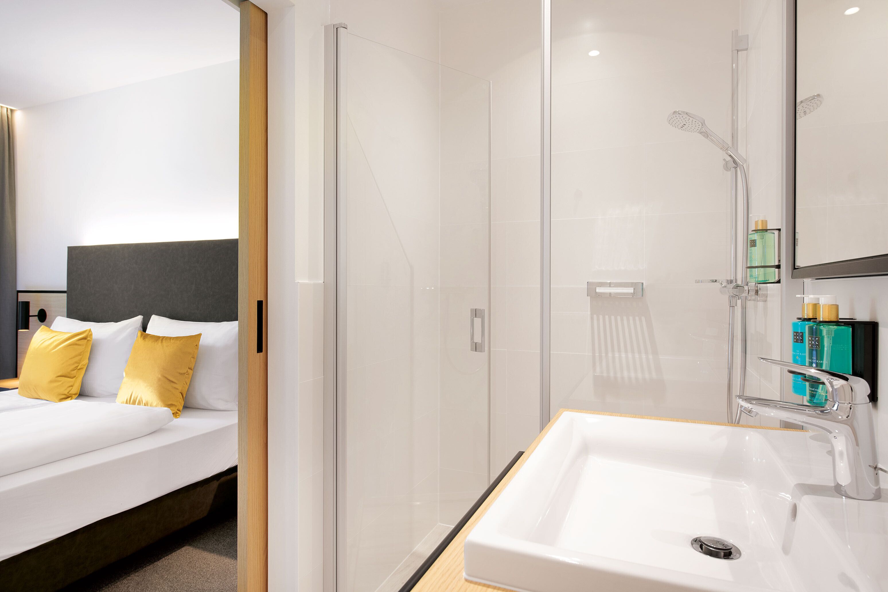 Una habitación de hotel moderna con una cama grande, cabina de ducha de vidrio y un lavamanos blanco con espejo.