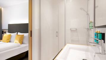 A modern hotel room with a large bed, a glass shower enclosure, and a white sink with a mirror.