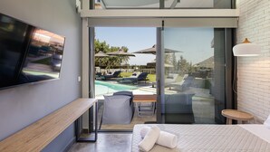A modern hotel room with a large bed, a flat-screen TV, a wooden bench, and a glass door leading to a pool area with lounge chairs and umbrellas. - Sauna, heated pool, kids pool, close to the beach & shops! (Rethymno)