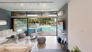 A modern living room with a large glass door leading to a pool area, a sofa, ottomans, and a small table. - Sauna, heated pool, kids pool, close to the beach & shops! (Rethymno)