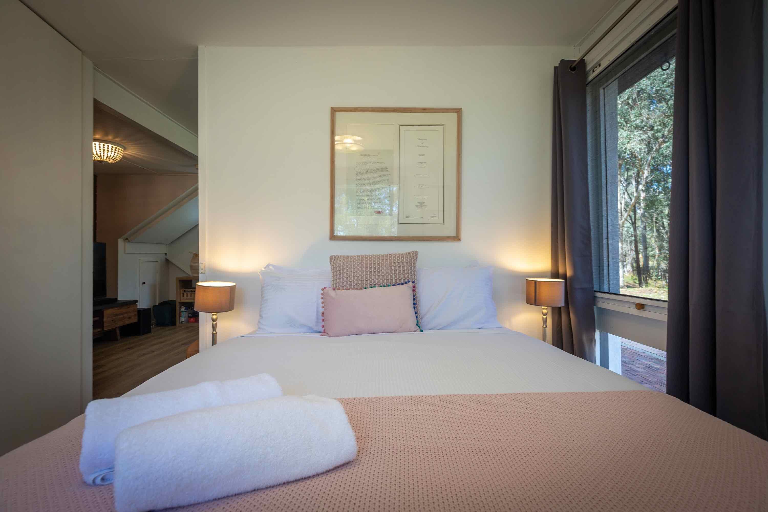 A neatly made bed with white linens and a pink blanket, a framed menu on the wall, and a window with curtains.