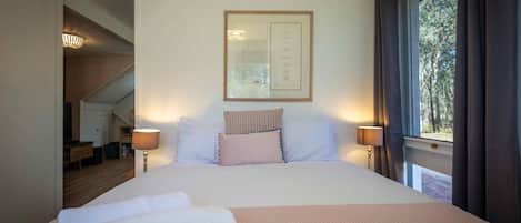 A neatly made bed with white linens and a pink blanket, a framed menu on the wall, and a window with curtains.