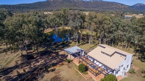 A single-story house with a large deck, surrounded by trees and a lake, with a hill in the background. - Marlu Station - Secluded family getaway in Broke (Broke)