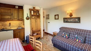 A compact living space with a kitchenette, a wooden cabinet, a sofa with patterned cushions, and a small bedside table. - Cozy cabin studio near ski lift (Combloux)