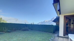 A well-maintained lawn with a hedge and a stone border, a patio area with a table and chairs, and a mountain view in the background. - Cozy cabin studio near ski lift (Combloux)