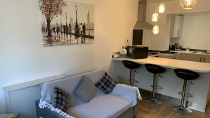 A modern kitchen with a central island, bar stools, and a painting of a cityscape. - Stylish and Immaculate 2-bed Apartment in Leigh (Leigh)