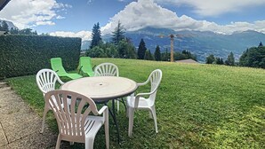 Eine Terrasse mit rundem Tisch und weißen Plastikstühlen, mit Blick auf eine Rasenfläche und eine Berglandschaft.