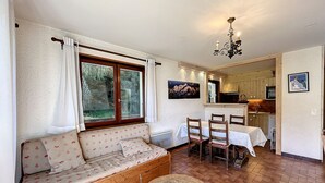 Ein gemütliches Wohnzimmer mit einem Holzdile, einem Esstisch mit Stühlen, einem kleinen runden Tisch und einer durchgehende Tür erkennbaren Küche.