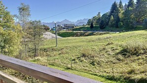 Ein grasbewachsenes Feld im Vordergrund, ein Gebäude mit geneigtem Dach im Hintergrund und eine Bergkette in der Ferne.