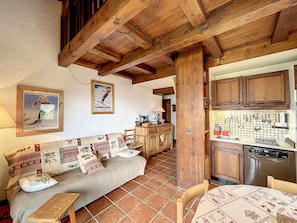 Ein gemütliches Zimmer mit Holzbalkendecke, einer Couch mit gemusterten Kissen, einem Esstisch mit Tischdecke und einer Küchenzeile mit Holdekor und Fliesenrückwand.