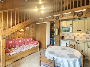 A cozy room with a wooden staircase, a sofa with patterned cushions, a dining table with a white tablecloth, and a kitchen area with wooden cabinets and a microwave.