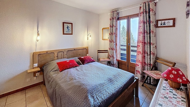 Une chambre à coucher avec un lit en bois, une table de chevet, une chaise, une table avec une lampe et une fenêtre avec des rideaux.