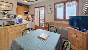 Une cuisine chaleureuse avec des meubles de rangement en bois, un four à micro-ondes, un évier et une table à manger avec des chaises. Il y a une horloge au mur et une fenêtre avec des rideaux.