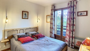 Une chambre à coucher avec un lit en bois, deux tables de chevet, une commode, une fenêtre avec des rideaux et une vue sur la verdure à l’extérieur.