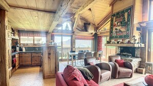 A rustic cabin interior with a wooden ceiling, a fireplace, a dining area, and a cozy seating area with a red sofa.