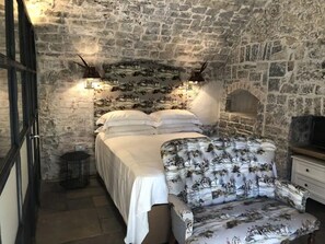 A bedroom with a stone wall, a bed with white linens, a patterned armchair, and a small table with a lamp.
