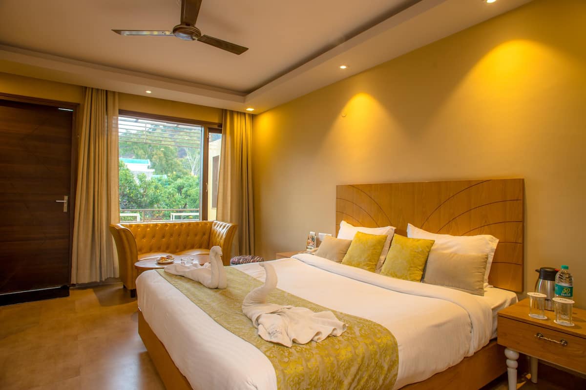 A hotel room with a large bed, a wooden headboard, a bedside table with a lamp, and a view of greenery outside.