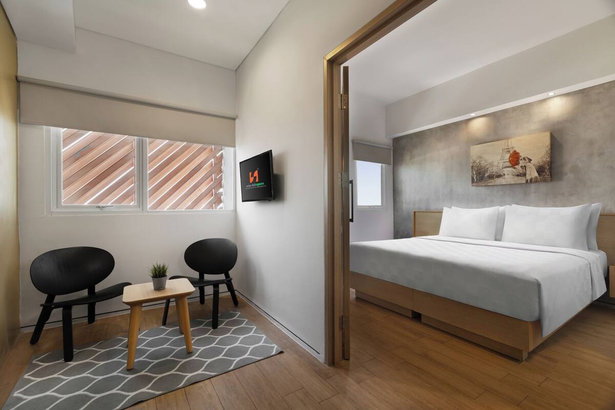 Ein modernes Schlafzimmer mit einem Bett, einem Fernseher, einem kleinen Tisch mit Stühlen und einem Fenster mit Jalousien.