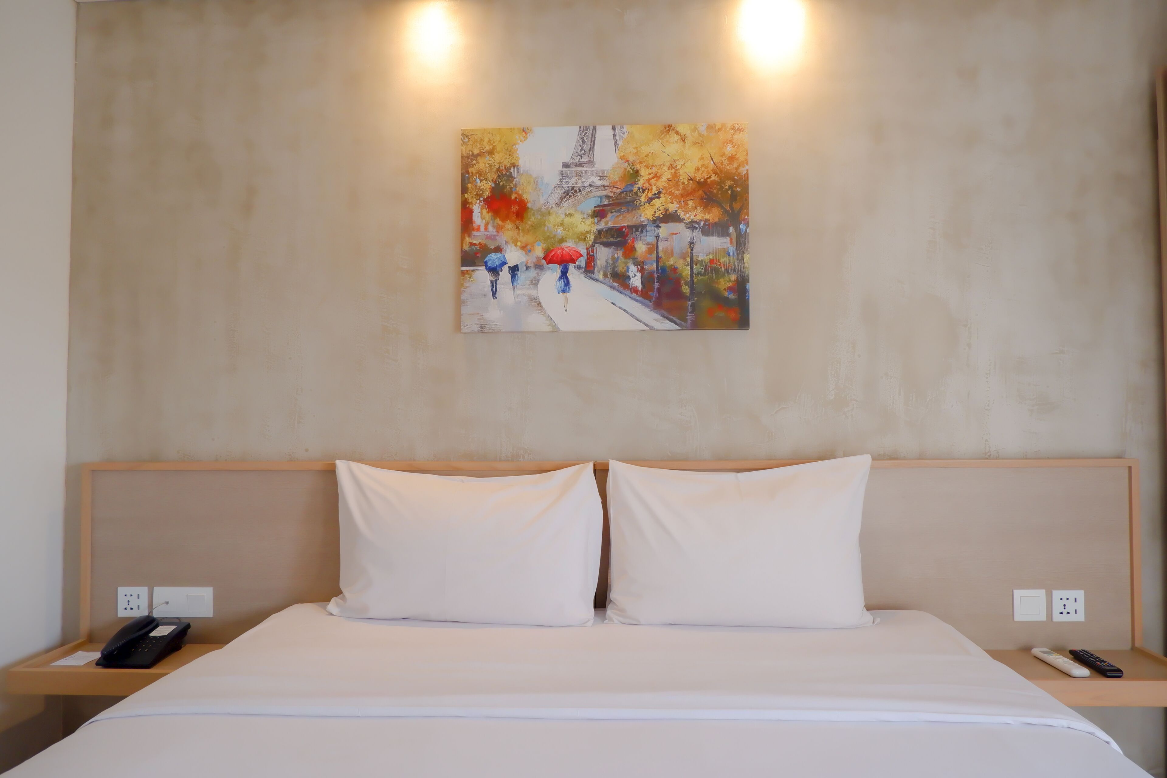A hotel room with a bed, two pillows, a nightstand with a phone, a remote control, and a painting of a Parisian street scene.