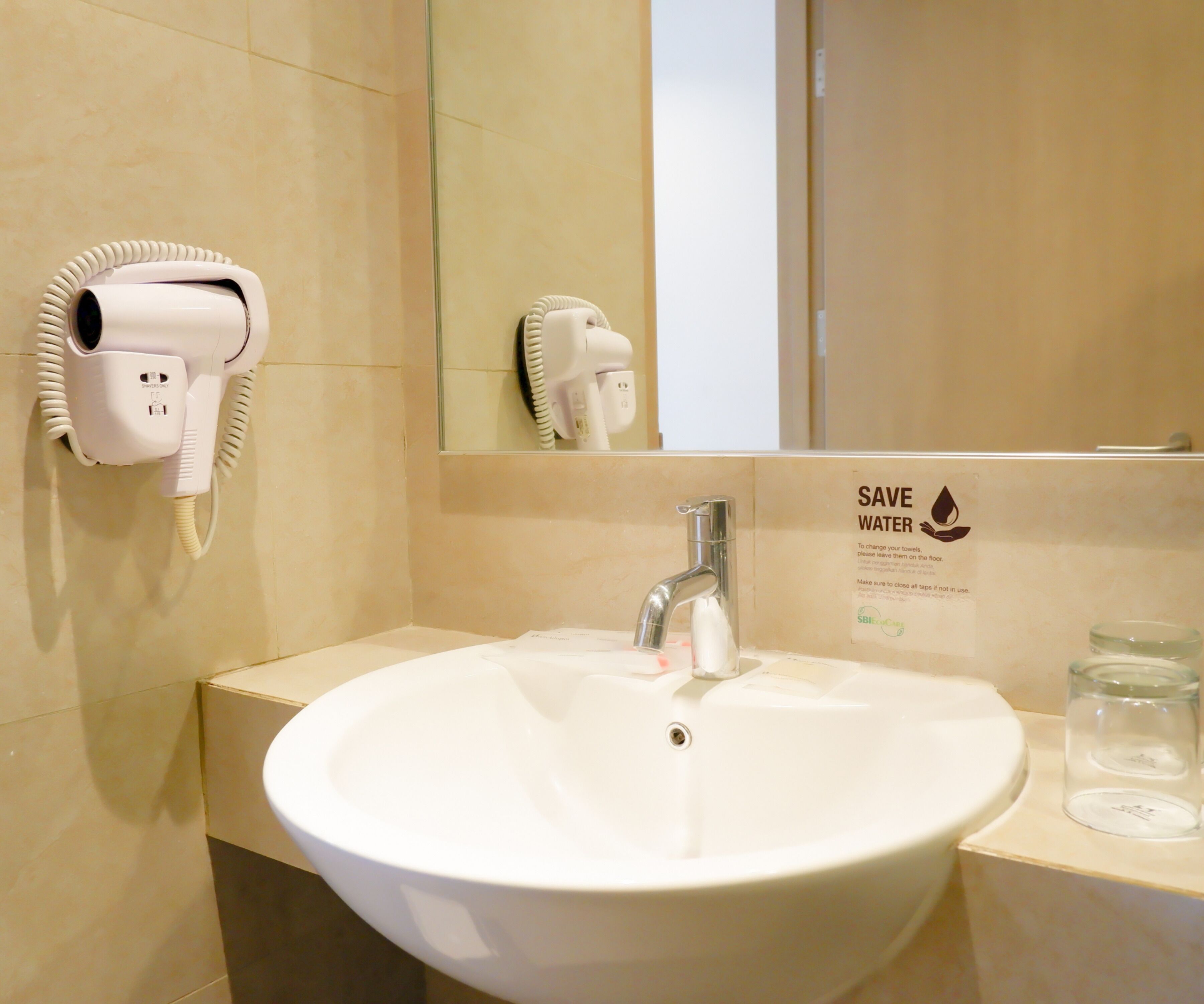 Une salle de bain avec un lavabo blanc, un miroir, un sèche-cheveux et un panneau indiquant « Économisez l’eau ».