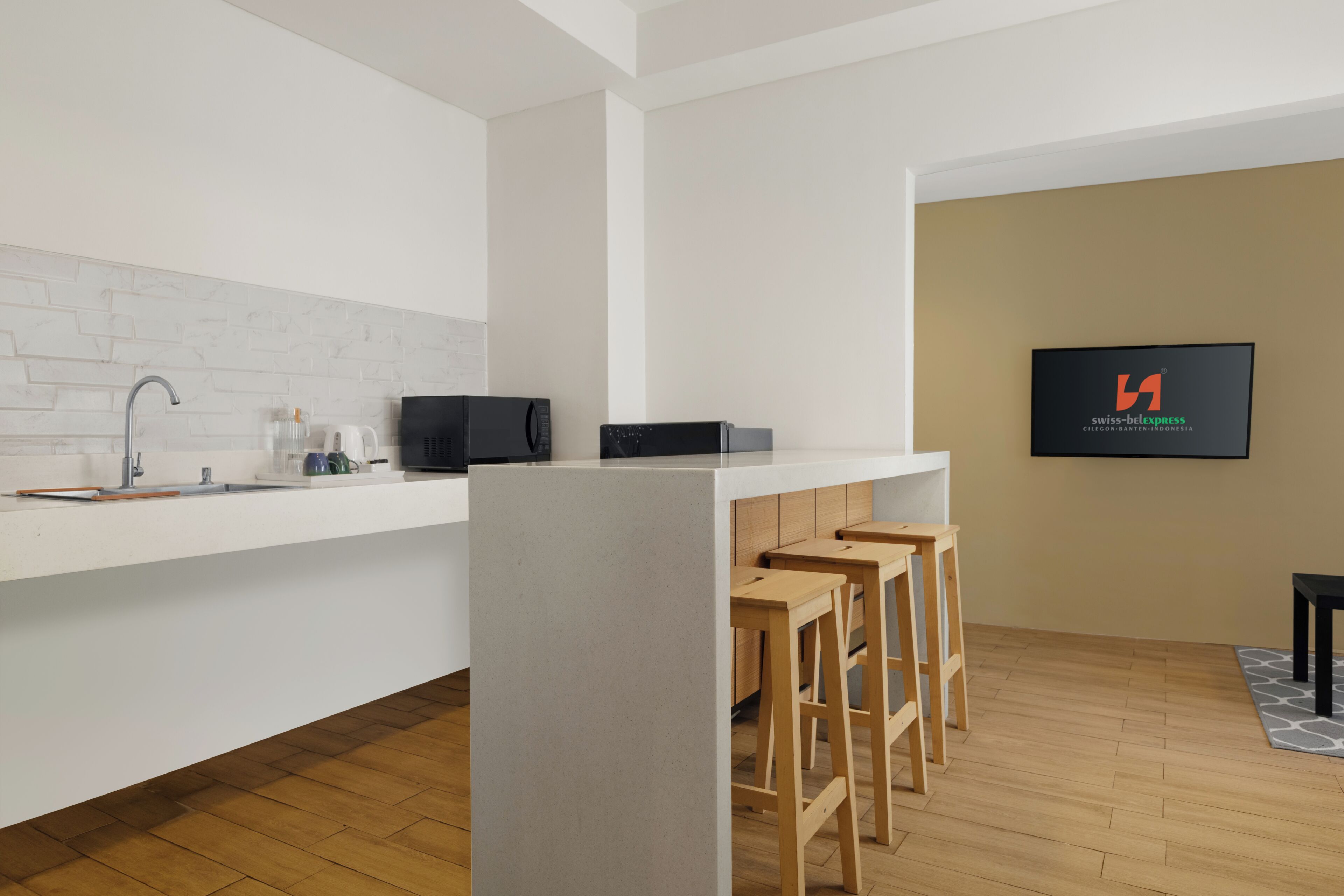 A modern kitchen with a white countertop, a sink, a microwave, and a refrigerator. There are wooden stools and a black table.