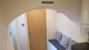 A compact room with a wooden door, a bench with patterned cushions, and a kitchen area with a black countertop. - 2-bed, Sleeps 4, Garden (London)