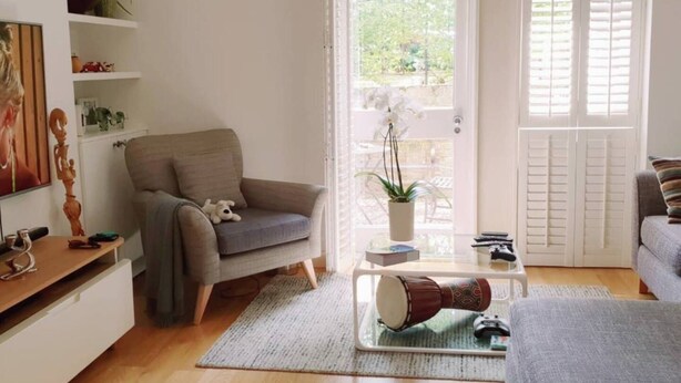 A living room with a grey sofa, a coffee table with a drum, a small table with a plant, and a window with white shutters. - 2-bed, Sleeps 4, Garden (London)