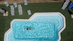 An aerial view of a swimming pool with a person swimming, surrounded by lounge chairs and a grassy area.