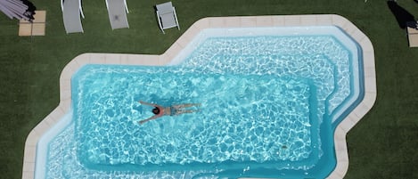 En vy från ovan av en swimmingpool där en person simmar, omgiven av solstolar och en gräsyta.