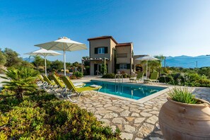 A pool area with lounge chairs and umbrellas, a stone patio, and a two-story building with a red-tiled roof. - Villa Helios, luxury holiday home, private pool, peaceful rural environment (Xirosterni, Crete)