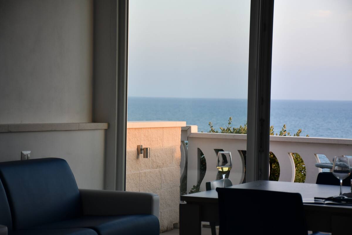 A balcony with a view of the sea, a table with a glass of wine, and a blue chair.