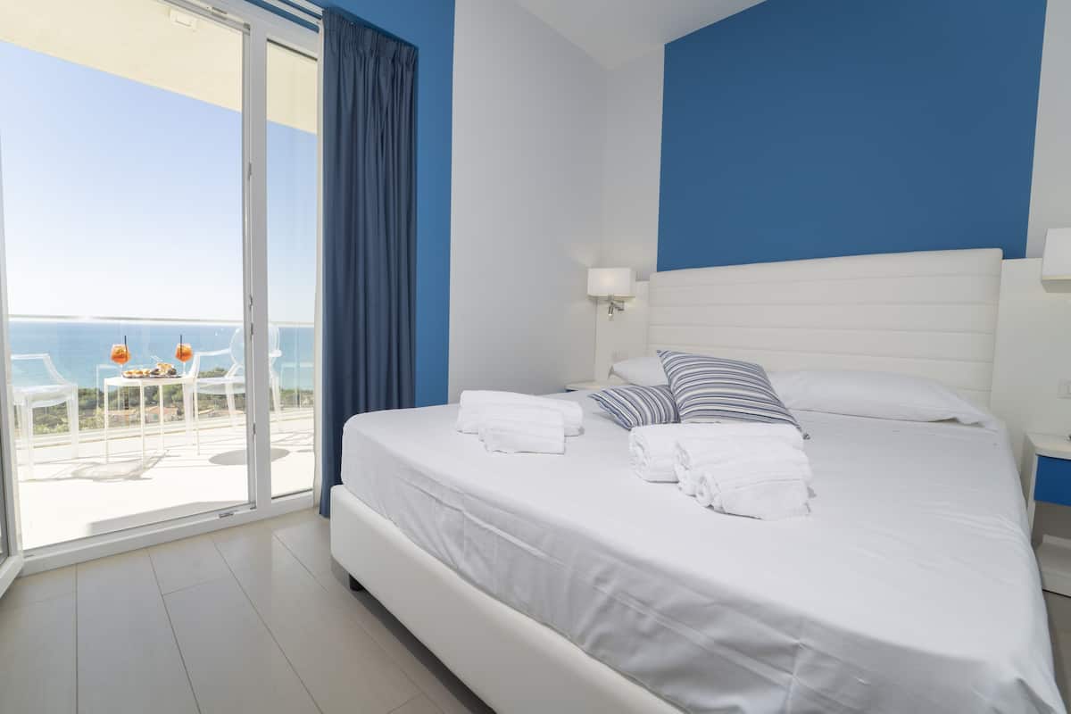 A hotel room with a bed, a nightstand, a lamp, a window with curtains, and a view of the sea.