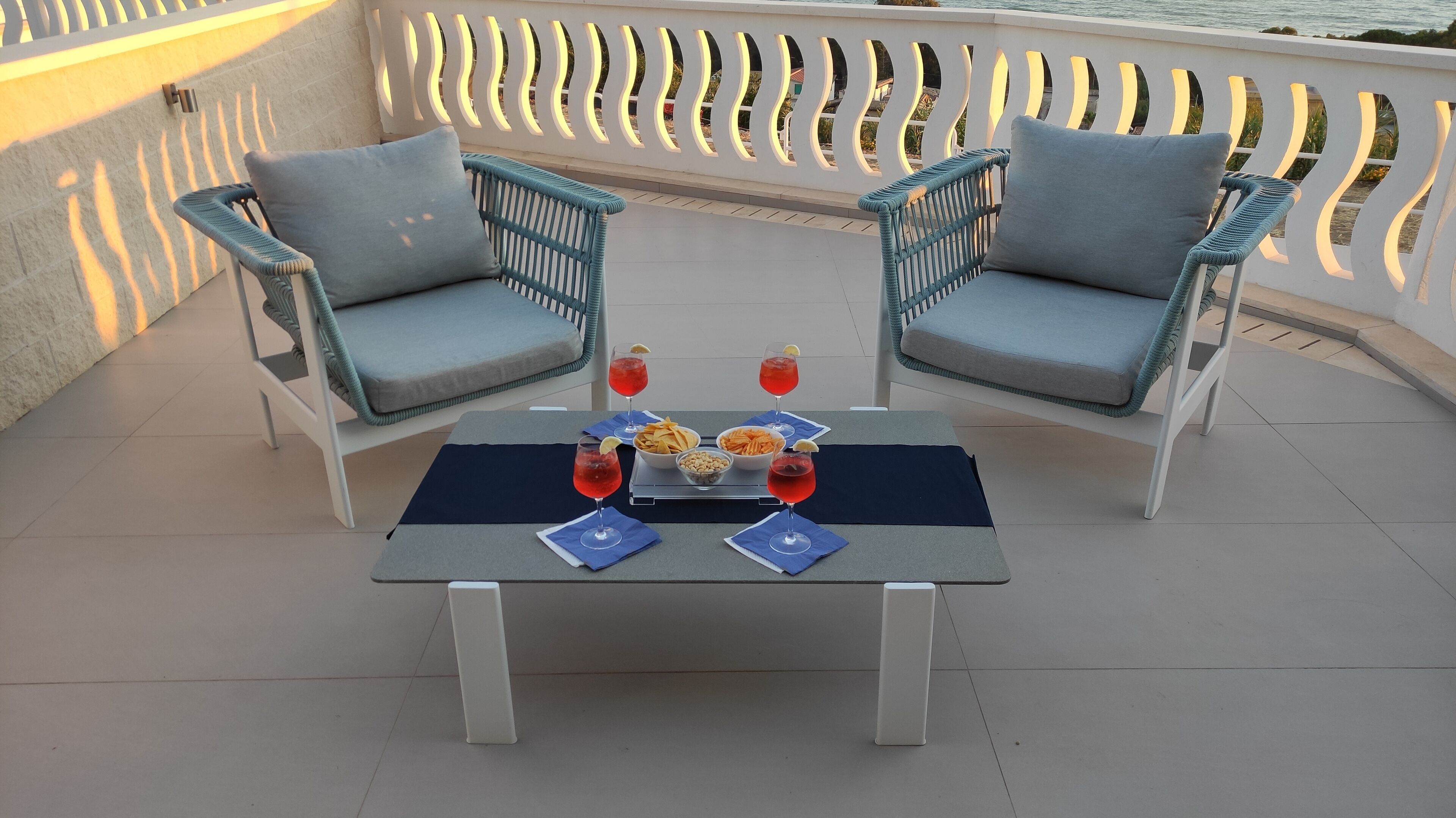 A terrace with two wicker chairs, a table with snacks and drinks, and a view of the sea.