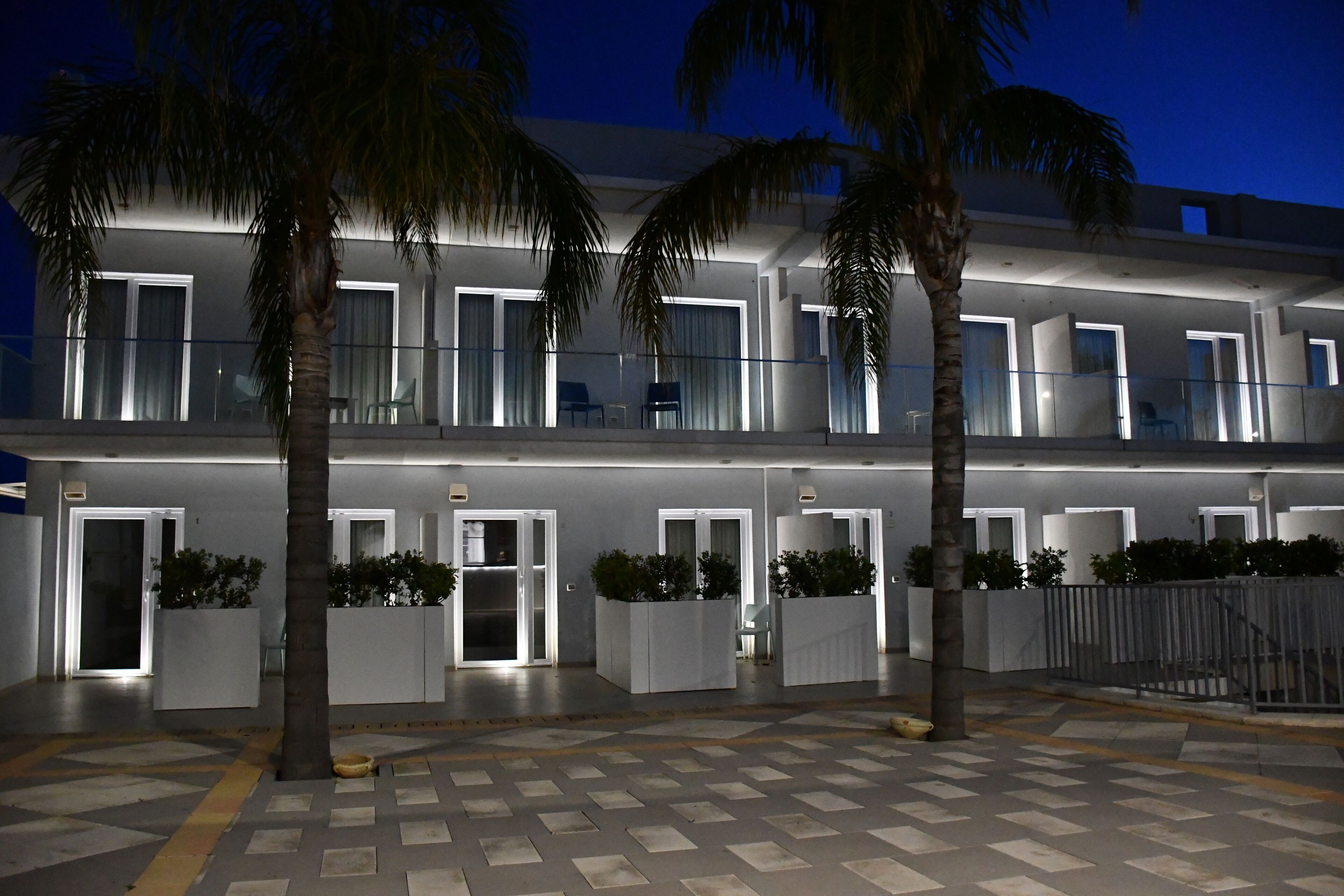 A modern hotel with a two-story structure, featuring balconies with glass railings, white exterior walls, and potted plants.