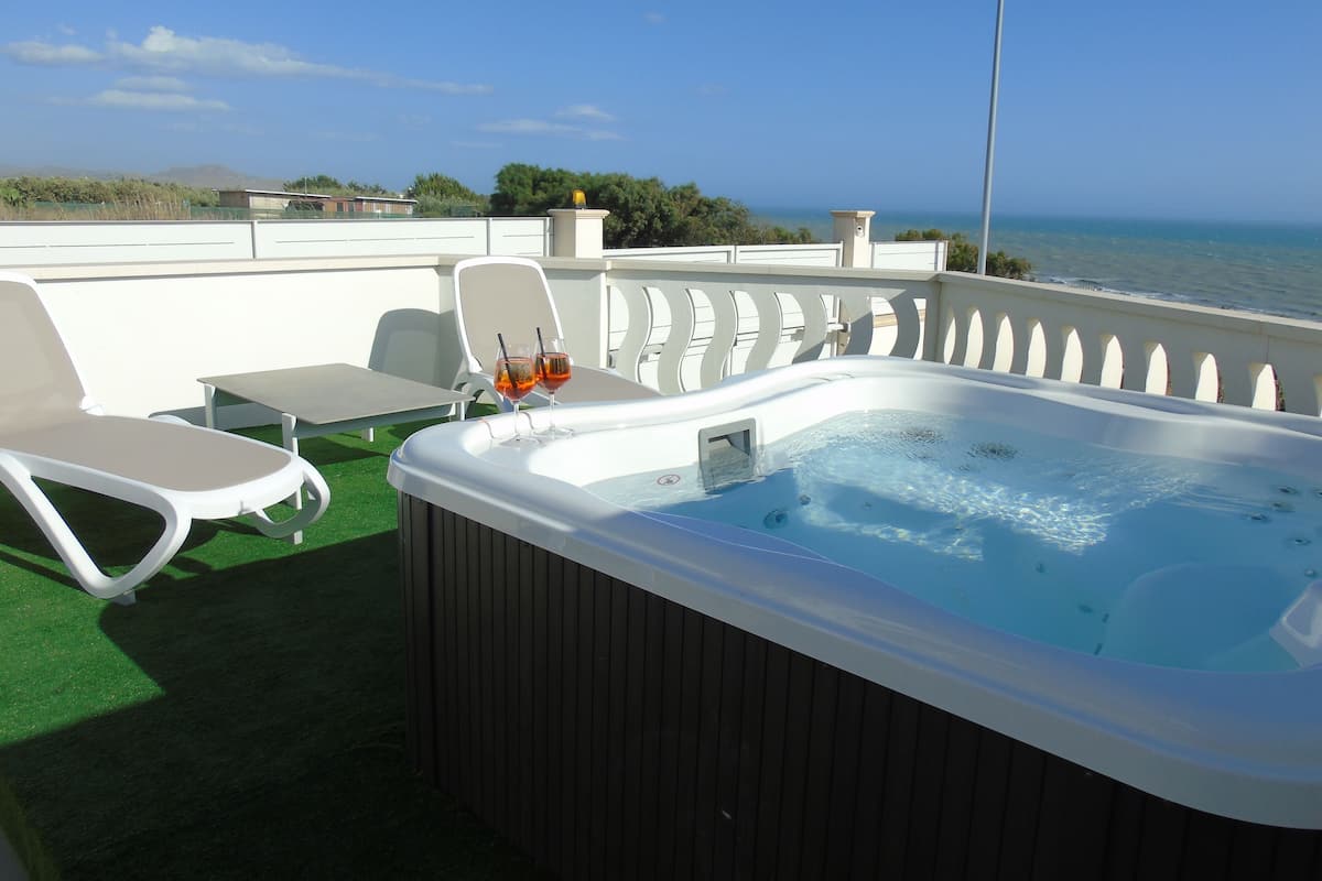 A hot tub with two glasses of drinks on a deck with lounge chairs and a table, overlooking the sea.