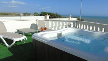 Un jacuzzi con dos vasos de bebidas en una terraza con sillones de descanso y una mesa, con vista al mar.
