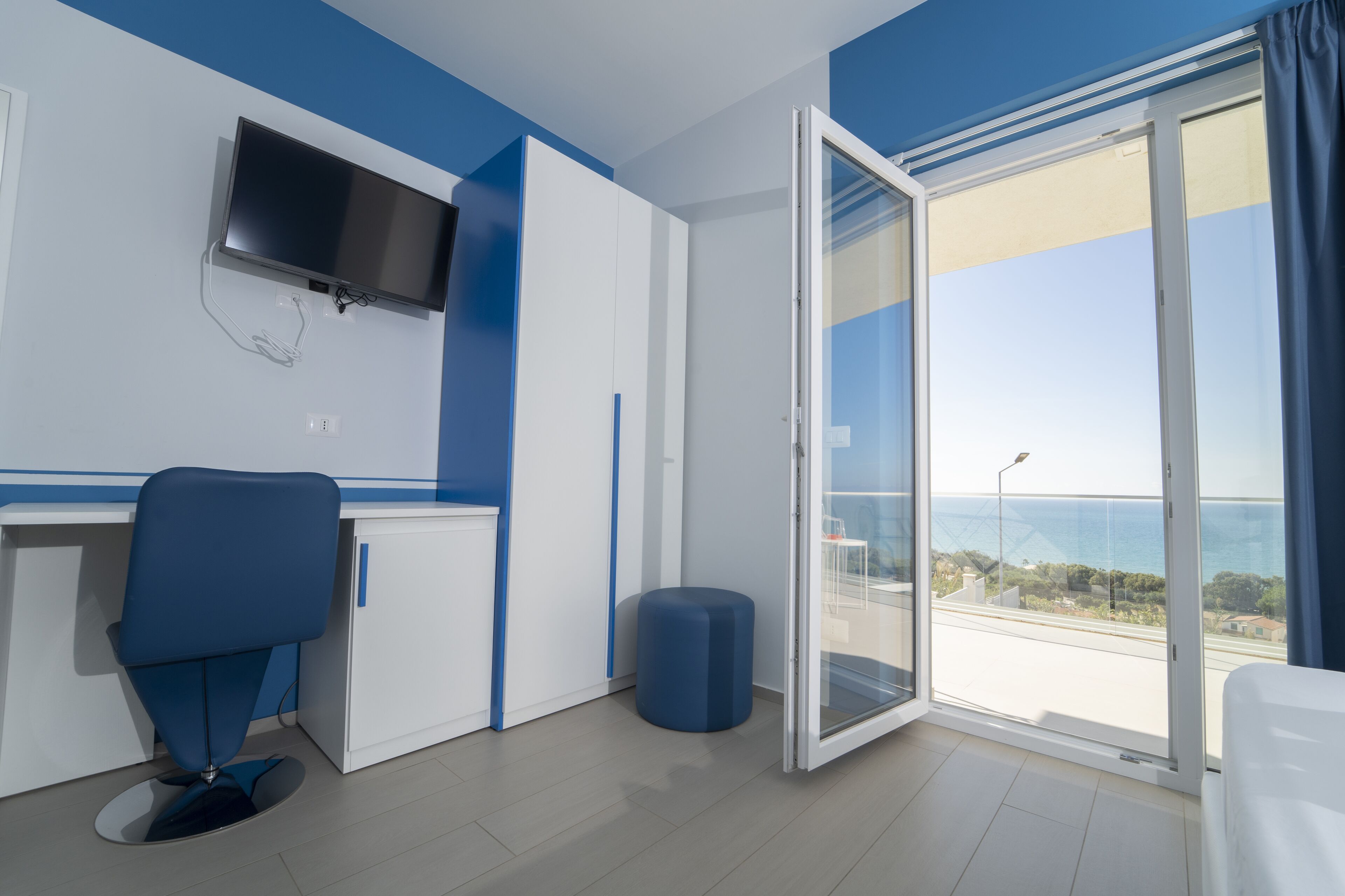 A modern hotel room with a desk, chair, and a large sliding glass door leading to a balcony with a view of the sea.