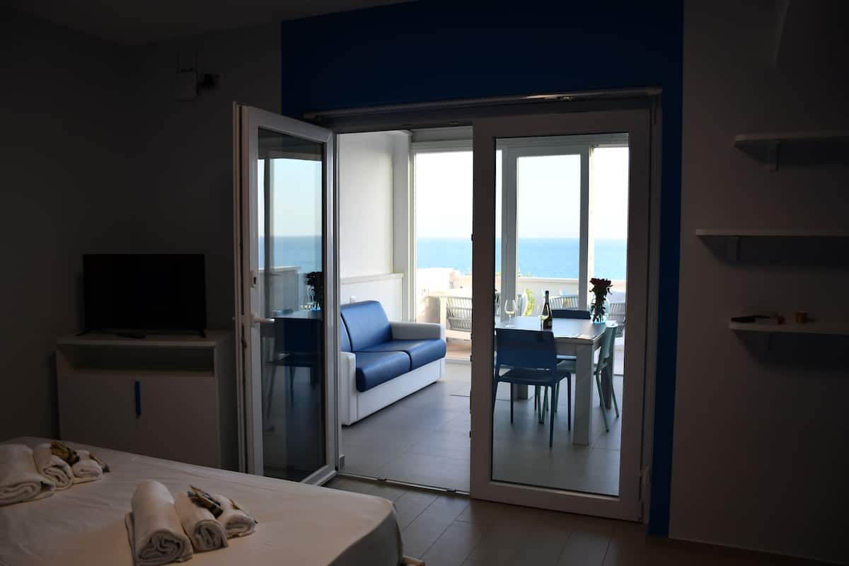 A hotel room with a bed, a TV, a sofa, and a balcony with a view of the sea.