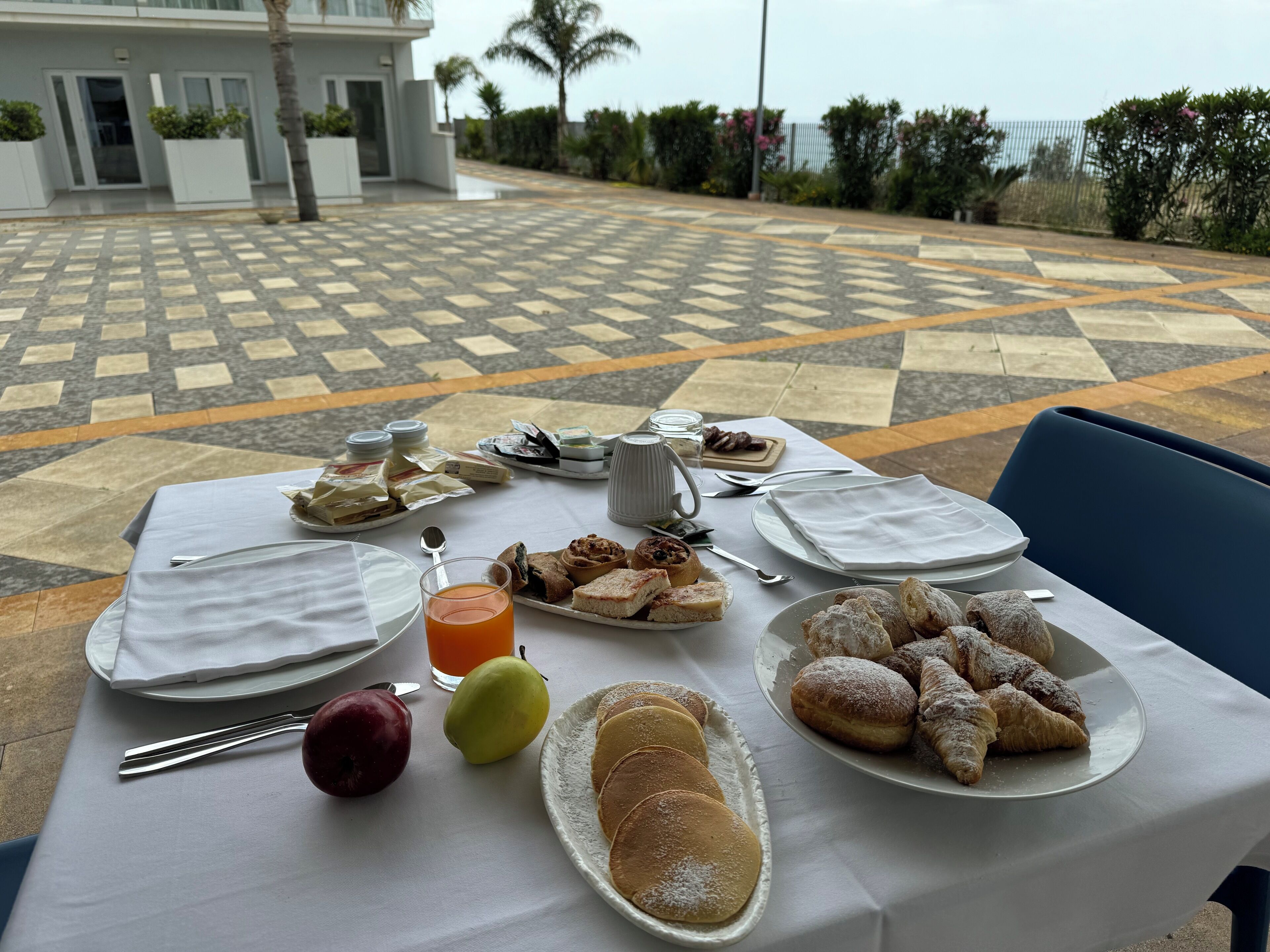 Une table dressée pour le petit-déjeuner, avec des viennoiseries, des fruits et une boisson, donnant sur un patio décoré de motifs.