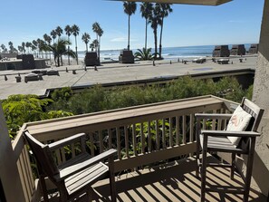 Un balcon avec un banc en bois et une table, donnant sur une plage bordée de palmiers et offrant une vue sur l’océan.