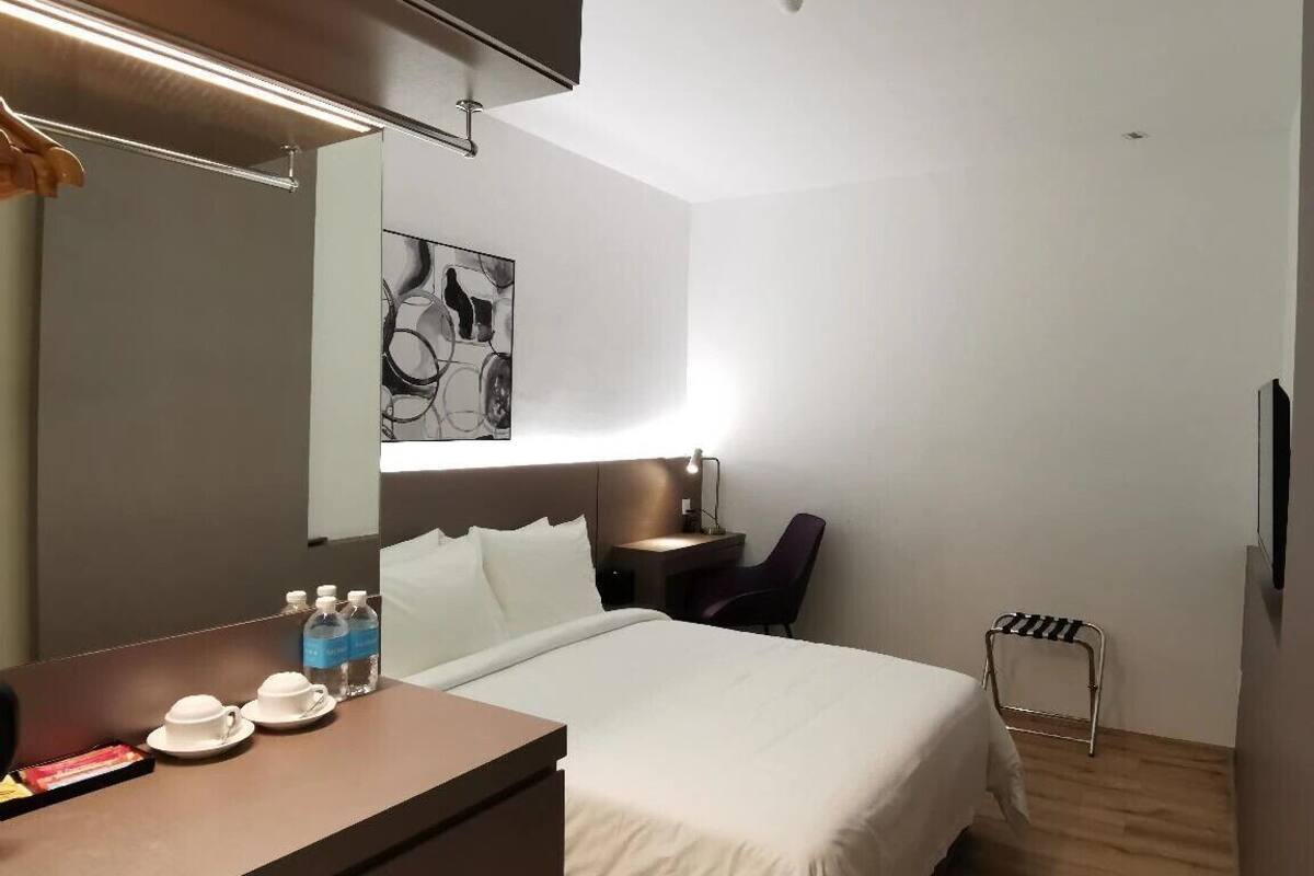 Ein modernes Hotelzimmer mit Bett, Nachttisch und Schreibtisch mit Stuhl. An der Wand befinden sich ein Spiegel, eine Lampe und ein kleines Kunstwerk.
