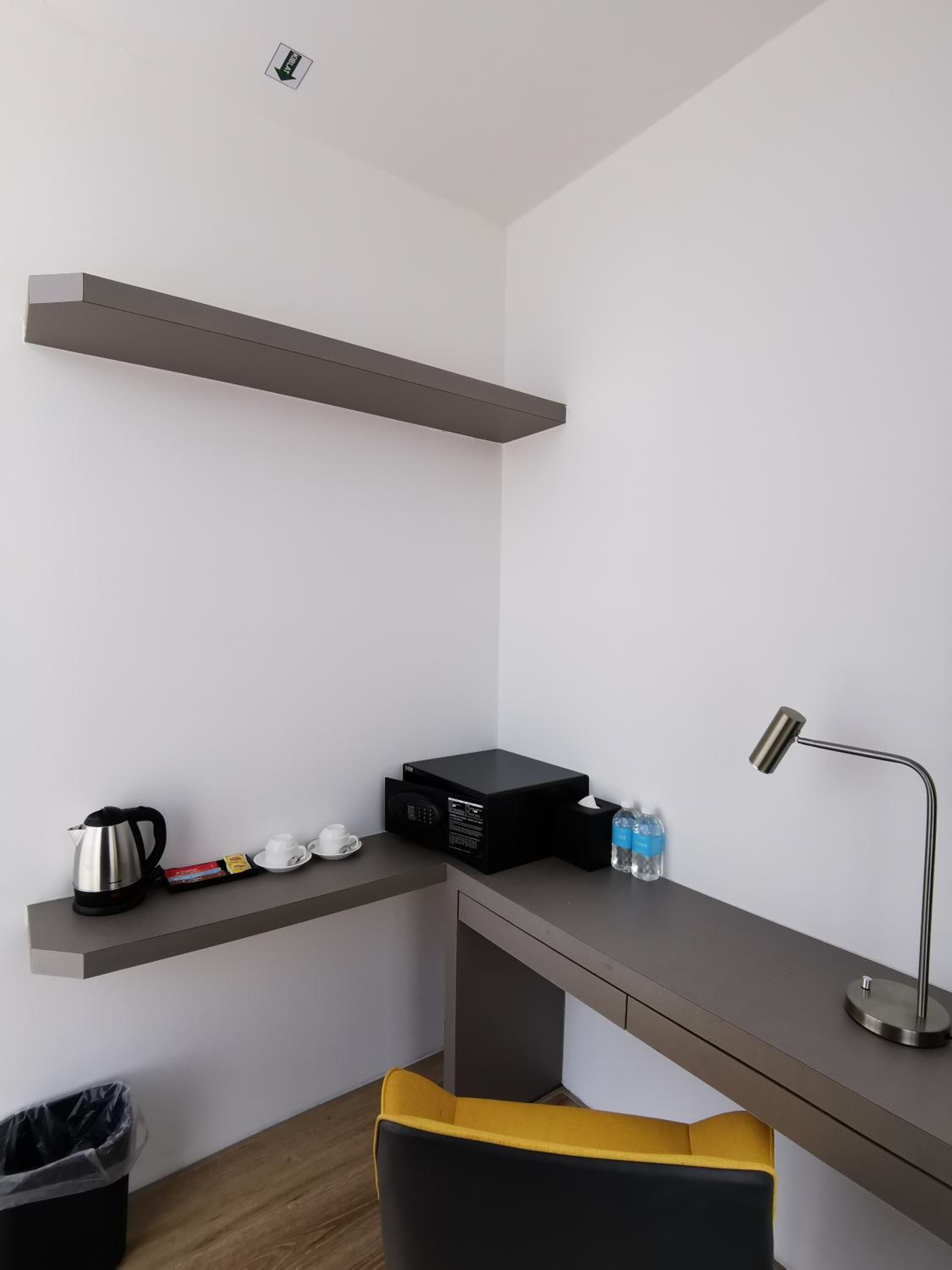 Une pièce moderne et minimaliste, avec un bureau gris, un appareil électronique noir, une bouilloire, une tasse, une bouteille et une chaise jaune.