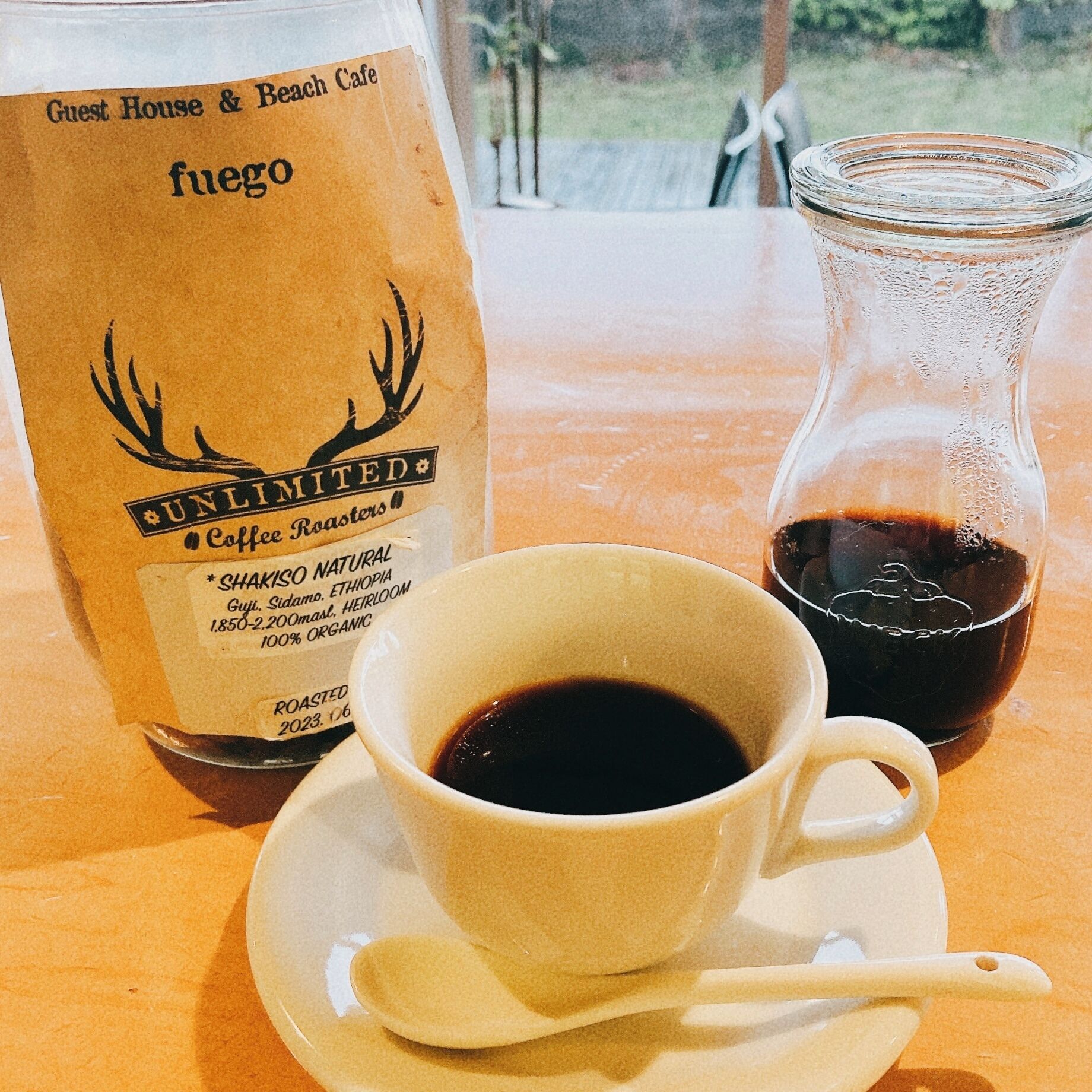A coffee cup on a saucer with a spoon, a glass bottle, and a paper bag labeled "UNLIMITED Coffee Roasters" with details about the coffee.