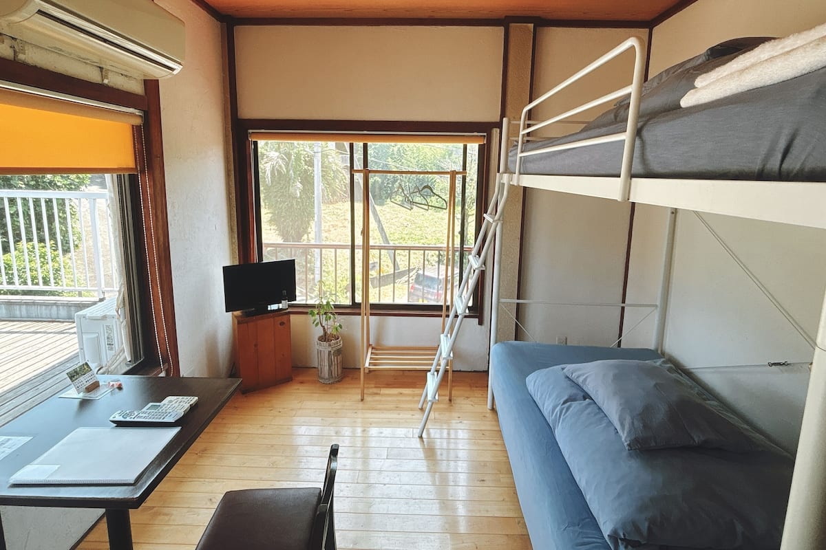 A compact room with bunk beds, a desk with a computer, a TV, and a window with a view of greenery.