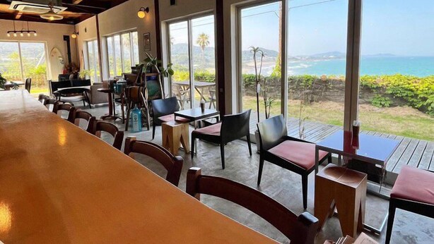 Un restaurante con una larga barra de madera y varias mesas con sillas. Amplias ventanas ofrecen vistas al océano y la vegetación.