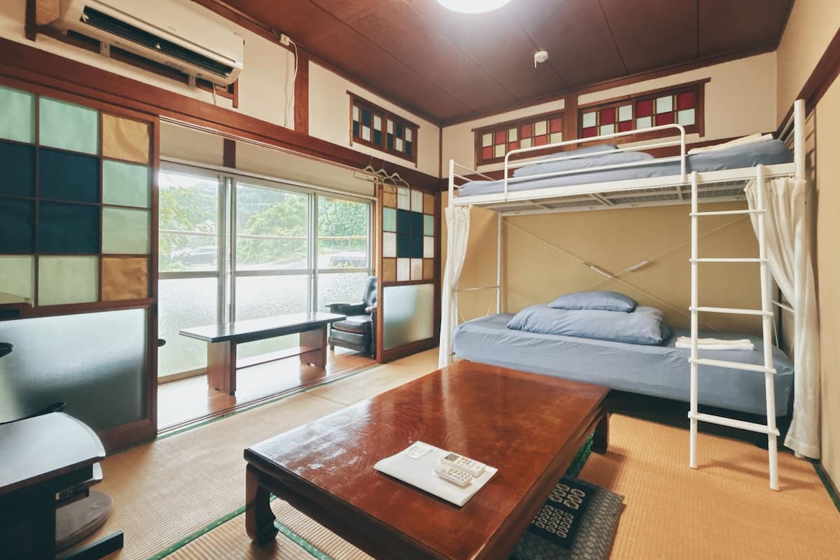 A room with a bunk bed, tatami flooring, a wooden table, and a view through large windows.