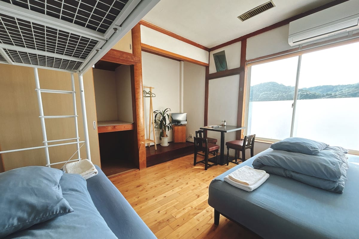 A room with a bunk bed, a desk with a chair, a small table, and a window with a view of a body of water.